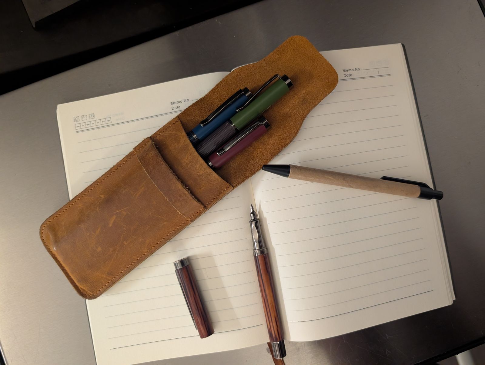 A leather pen roll lying open on a lined notebook, with a wooden fountain pen and a few other pens